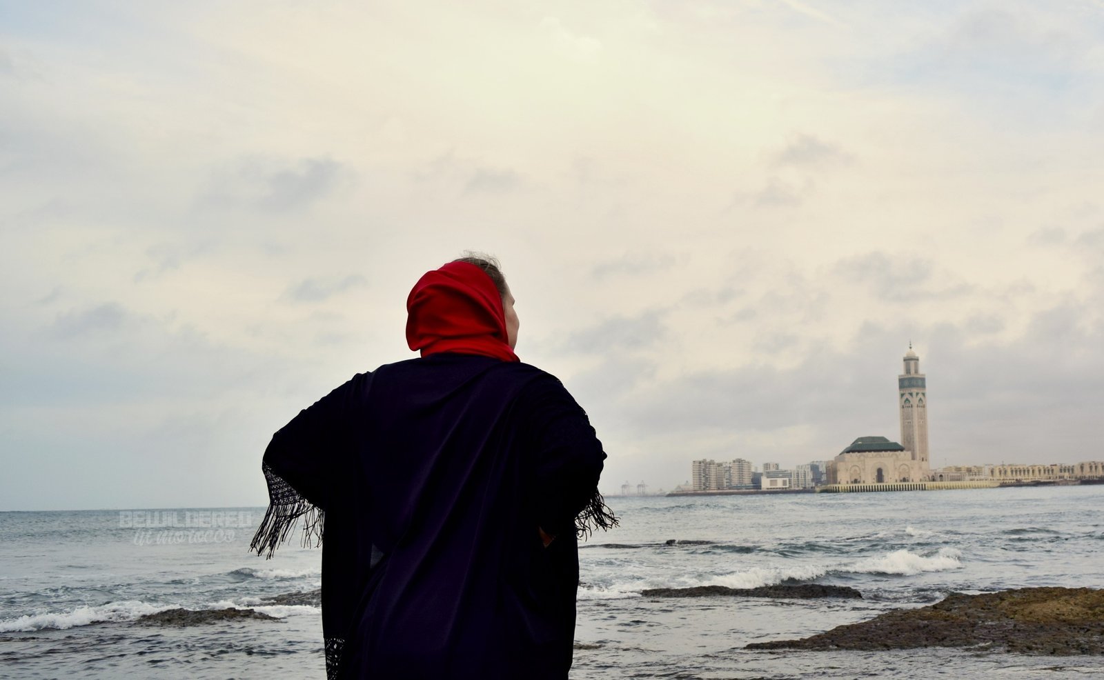 Woman in a red headscarf looking out at the Atlantic Ocean with the Hassan II Mosque minaret in the distance, Casablanca
